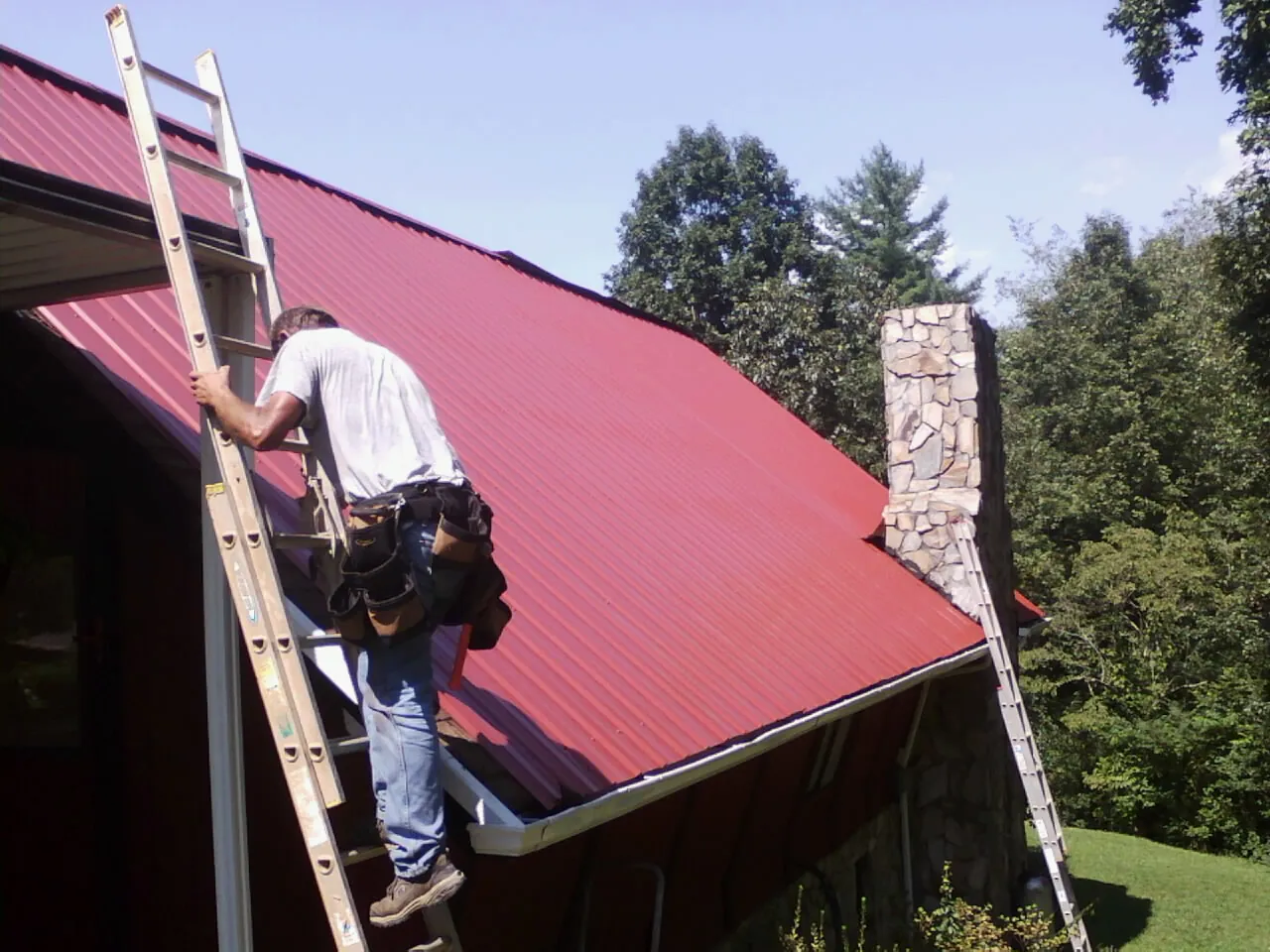 Roofer on a ladder beside a red metal roof and stone chimney — Soffit & Fascia Repair by Roofing Brothers in Soda Springs